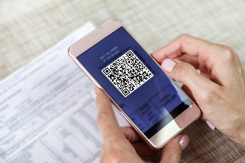A person scans a QR code displayed on a smartphone screen, with a paper document underneath on a table.