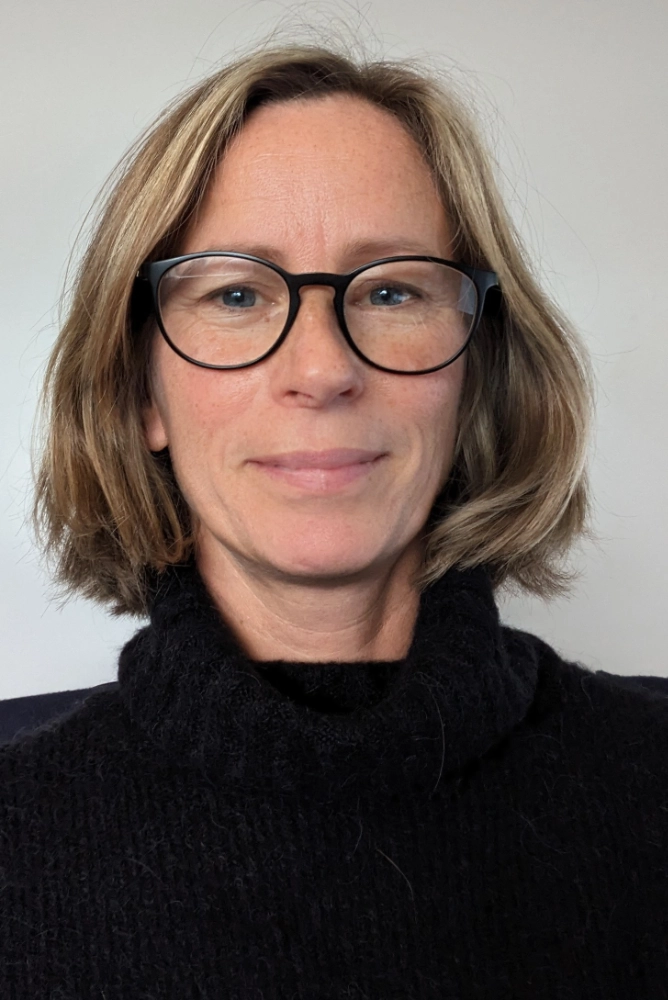 A person with shoulder-length light brown hair and glasses is wearing a black turtleneck sweater, facing the camera against a plain background.